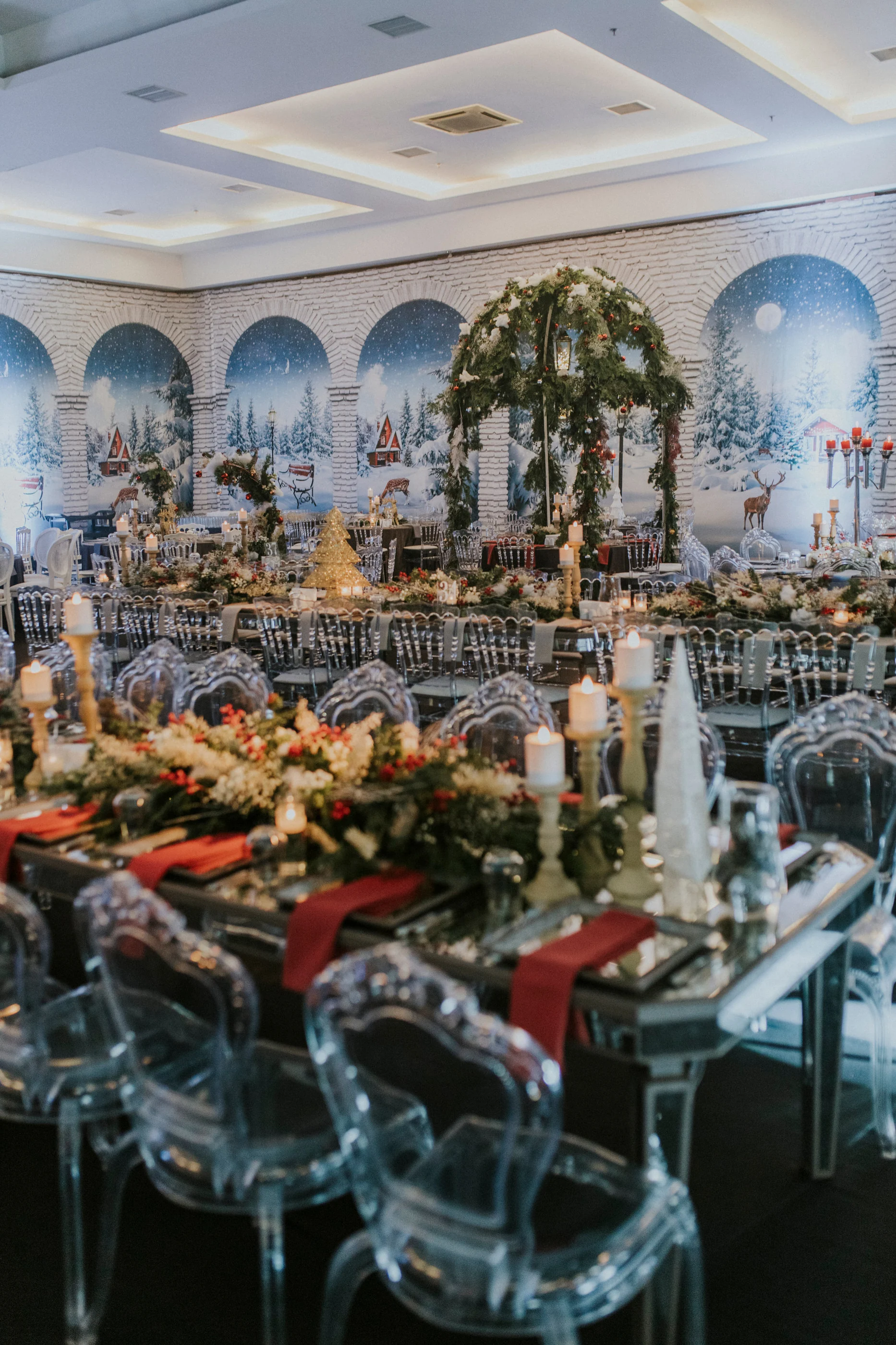 Mirrored tables with red napkins and clear chairs before arches painted with snowy forests