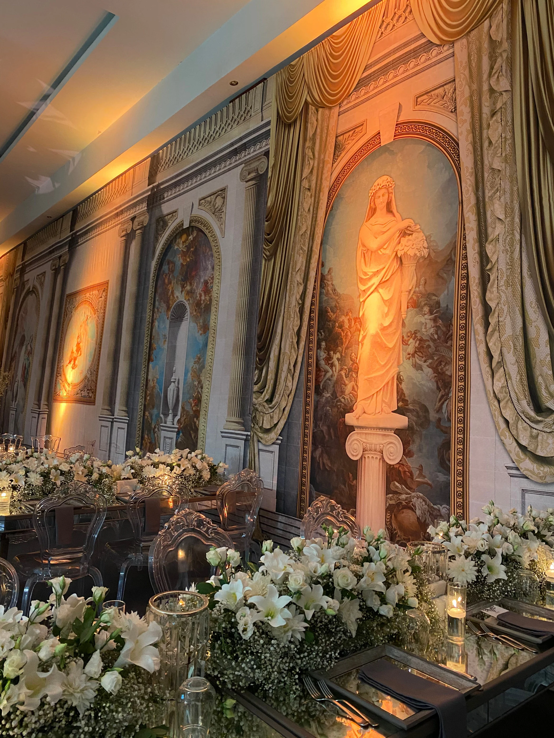 Illuminated statue on a column before a fresco wall, with a white lily table