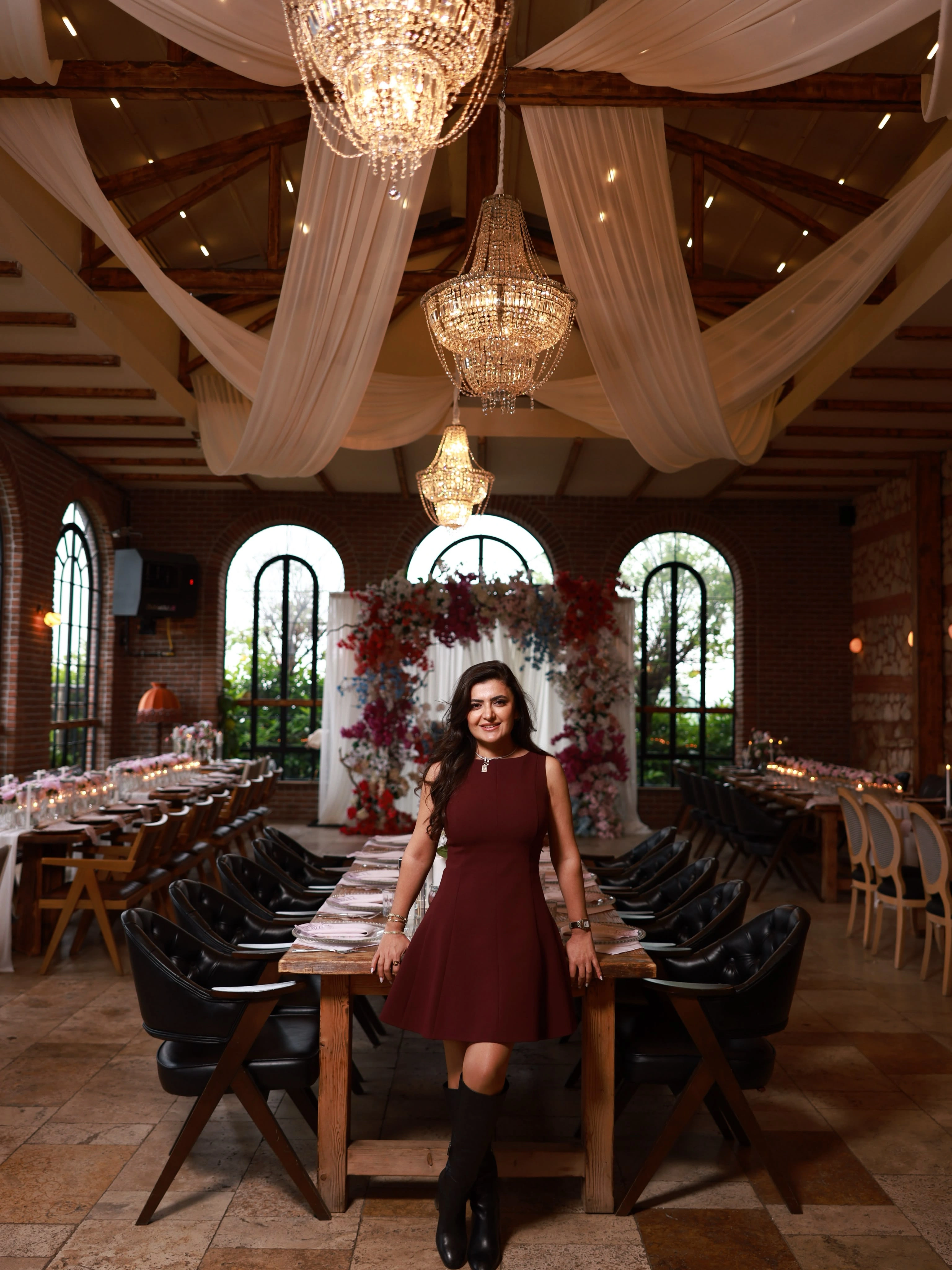 A woman in a burgundy dress at the head of a long table beneath draped tulle and crystal chandeliers