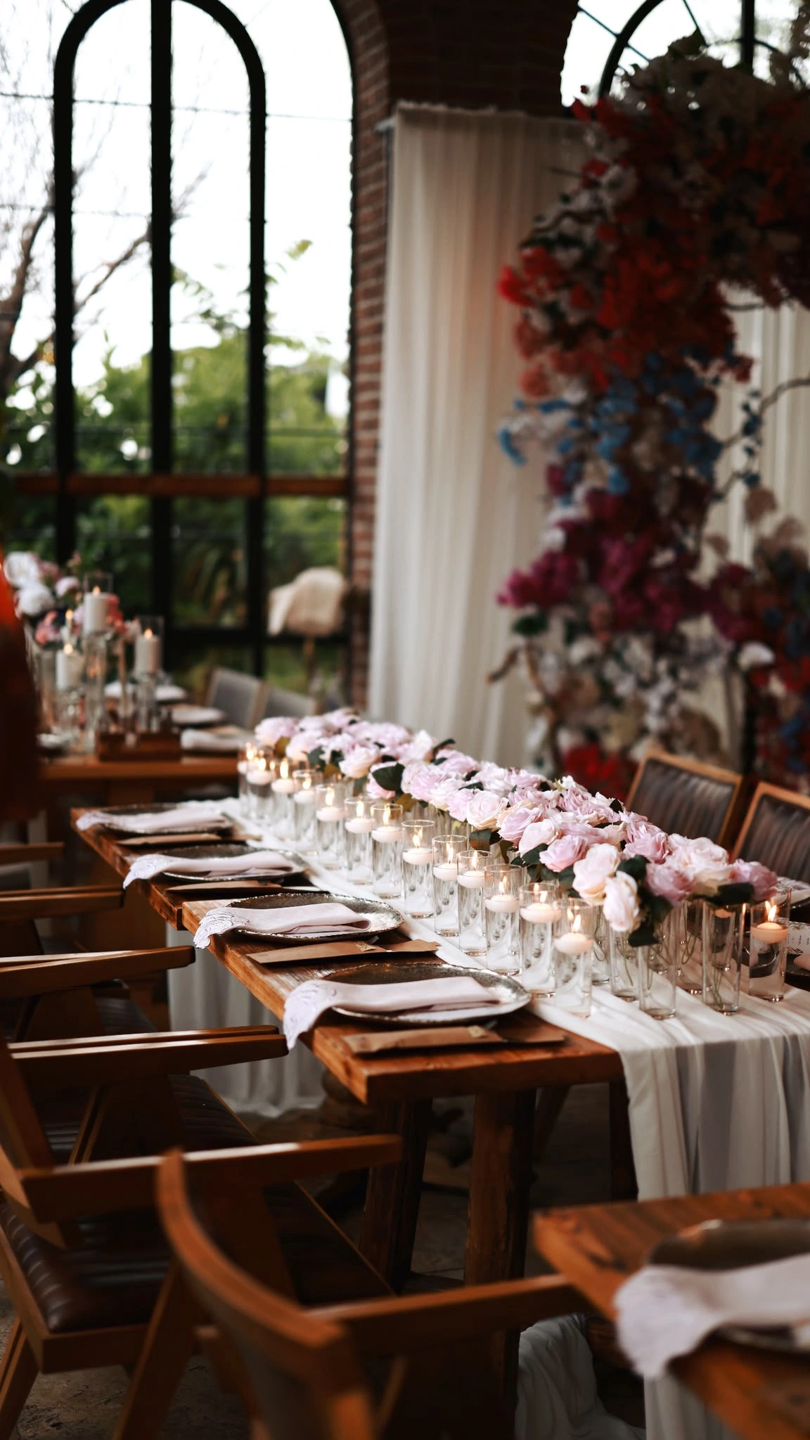 Table lined with pink roses beside a white sheer curtain and a deep red floral installation