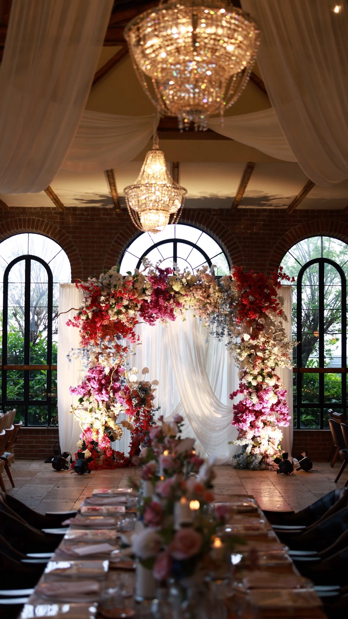 Floral arch of red and pink blooms with white drapes in front of three arched windows