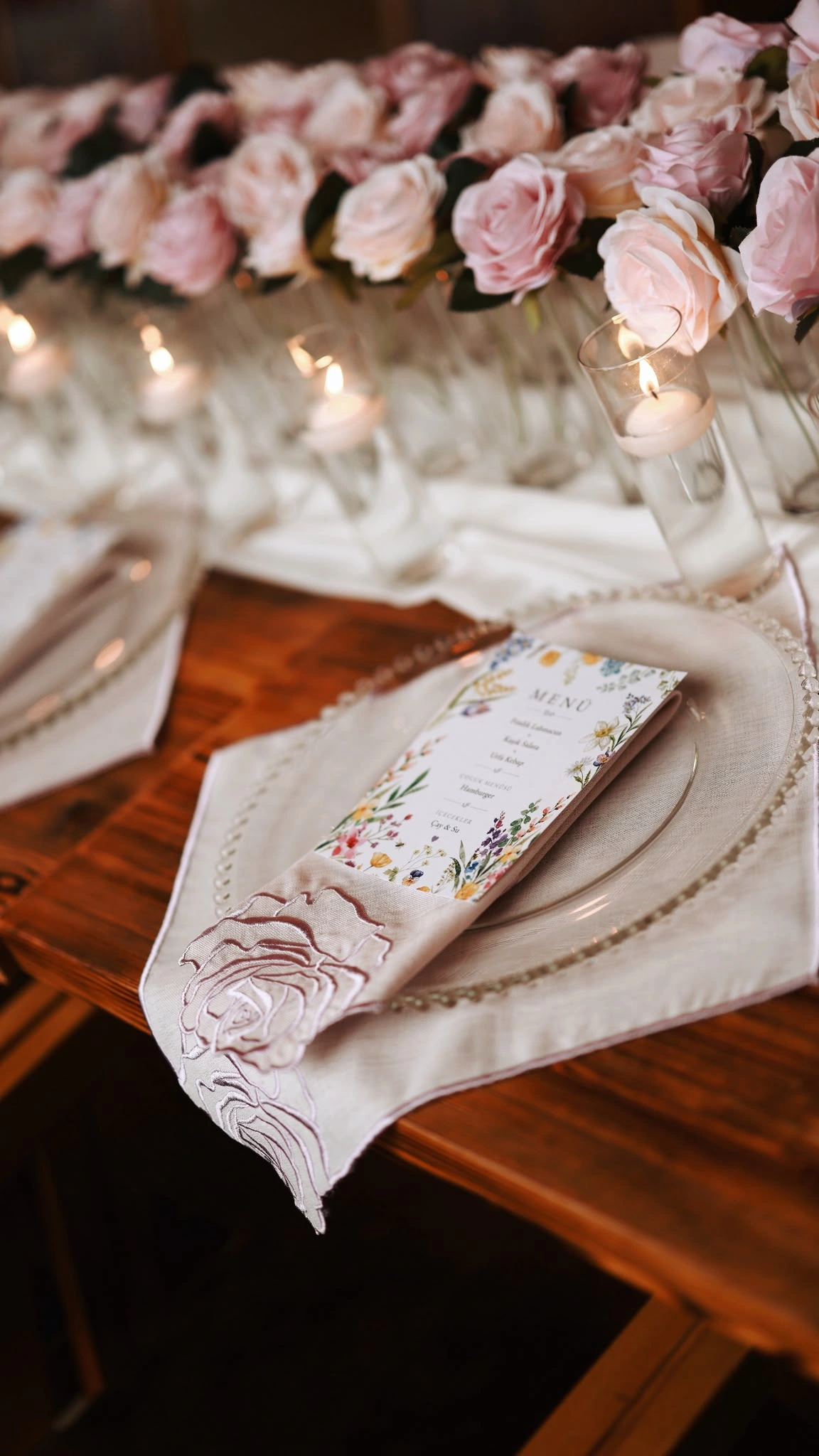 Floral-printed menu card on an embroidered napkin with pink roses and candles behind