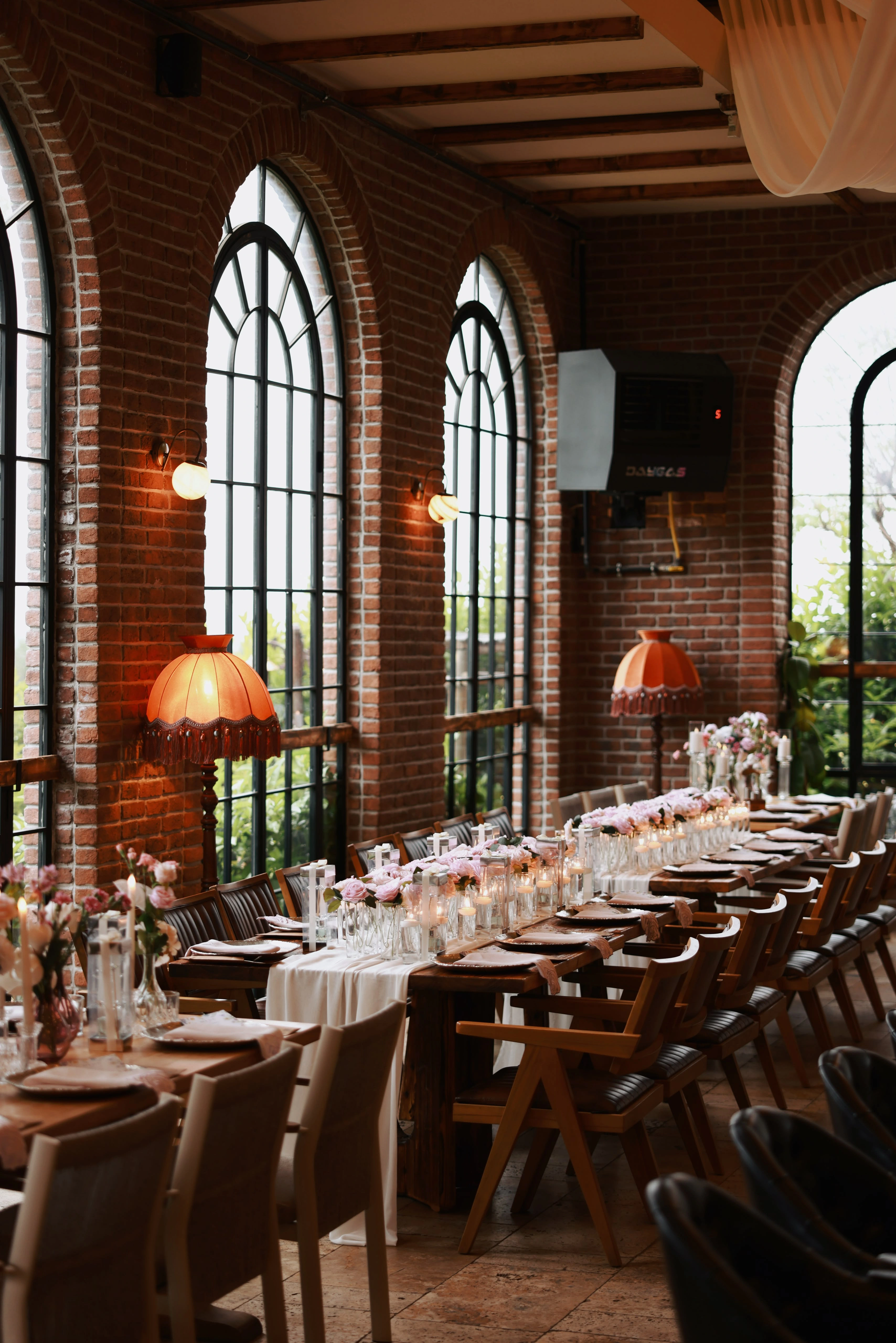 Overall view of the hall with tall arched windows, orange fringed sconces and brick walls