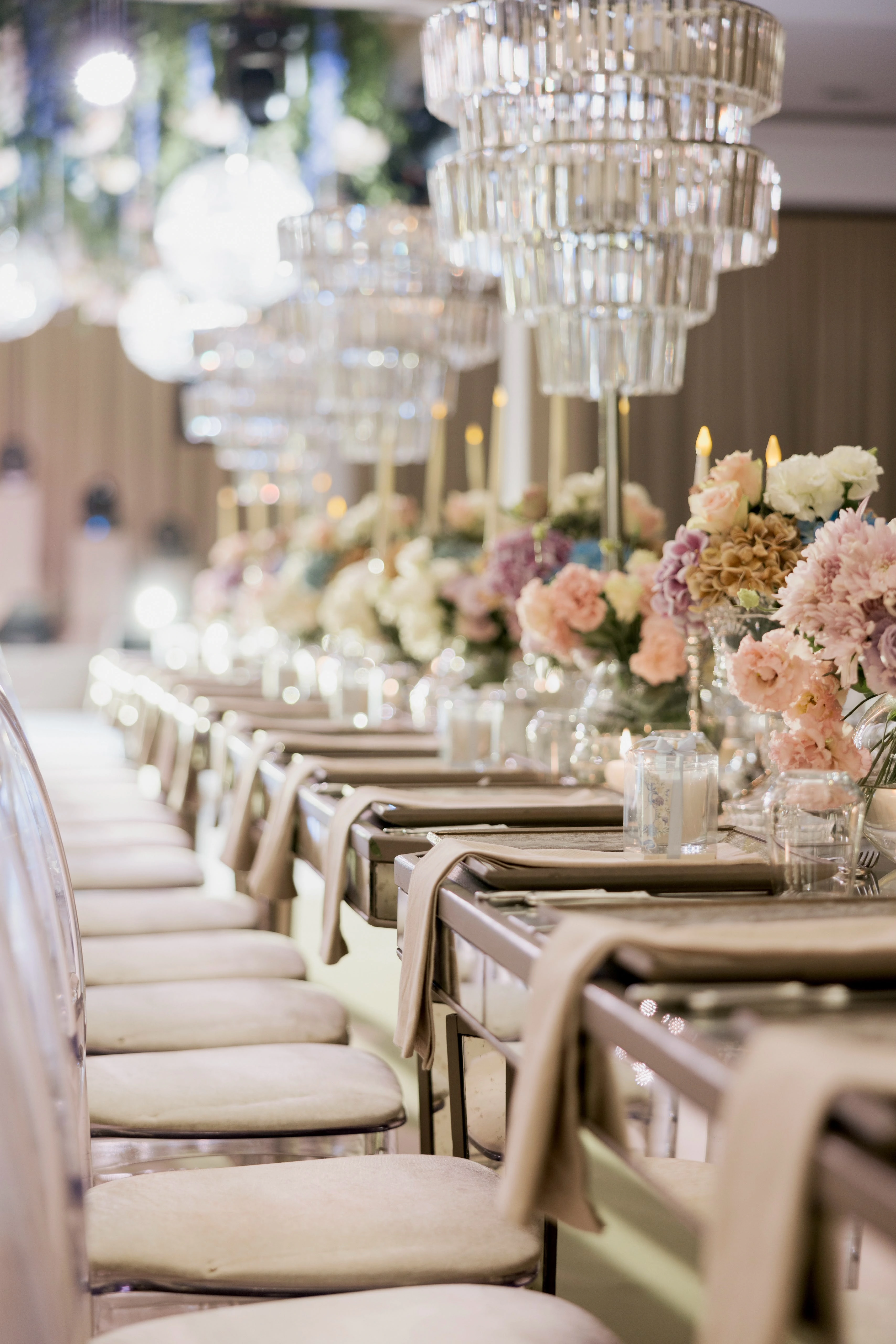 Perspective of tiered crystal centrepieces and pastel flowers along a long table with clear chairs