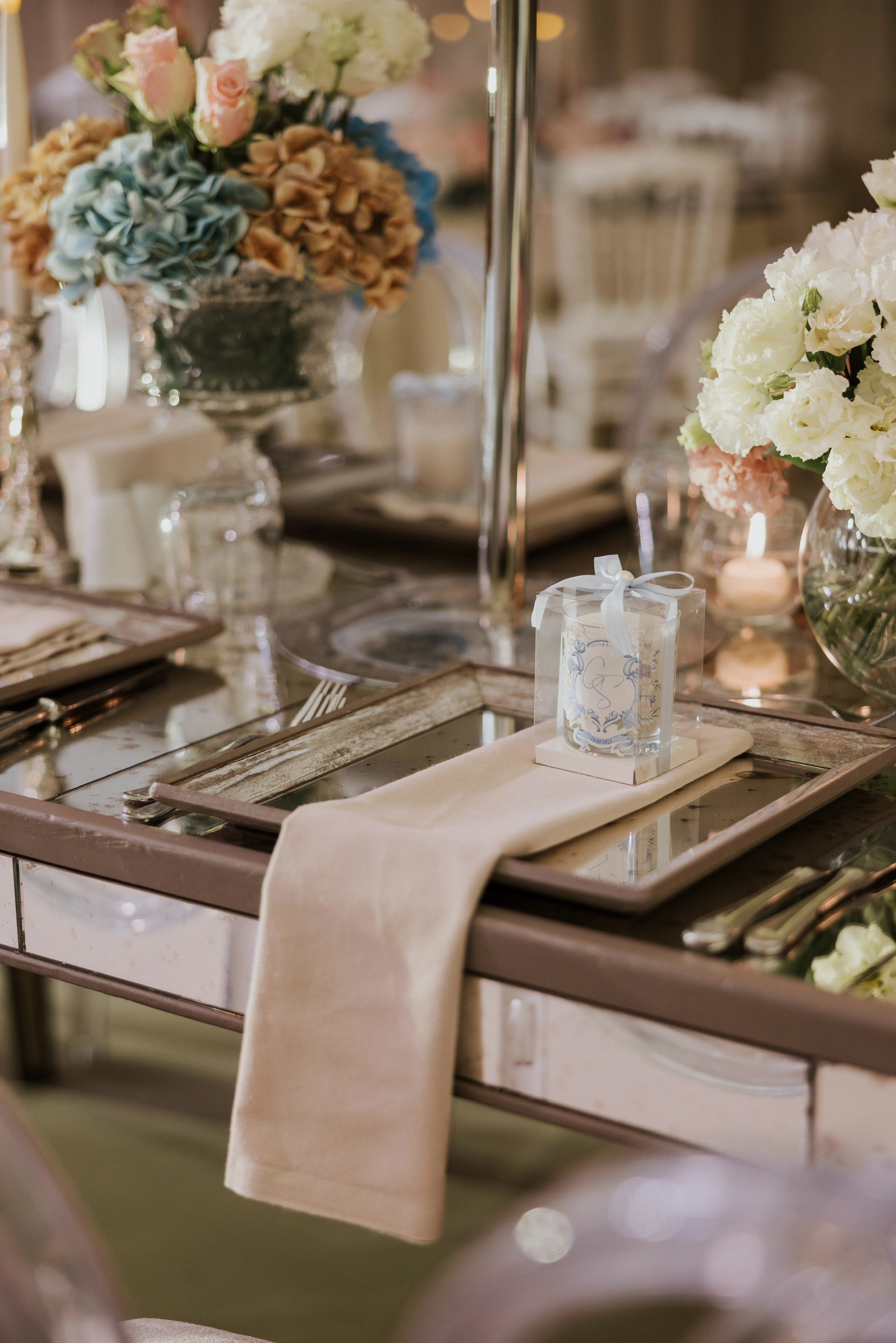 Close-up of a beige napkin, clear favour box and blue hydrangea arrangement on a mirrored table