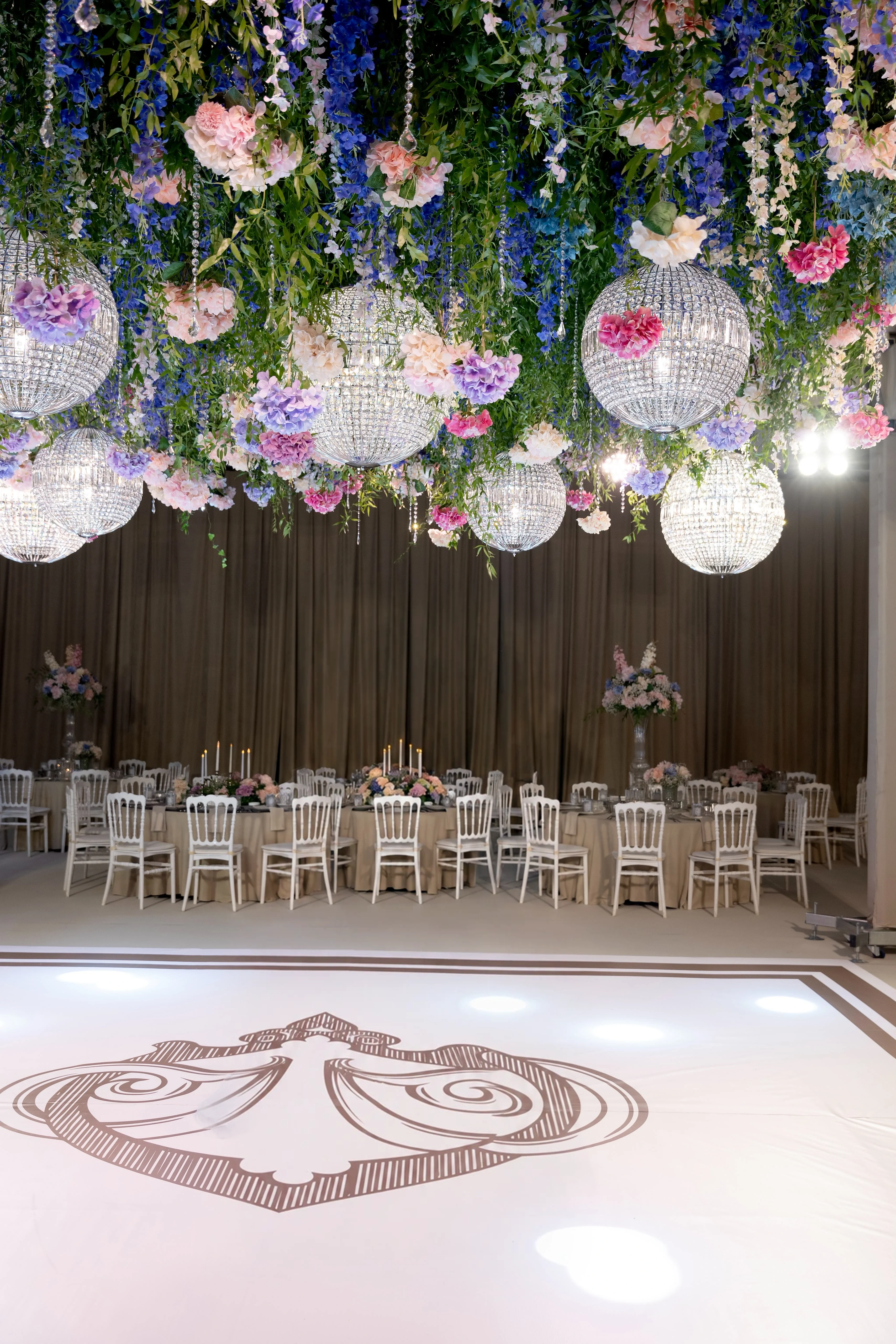 Crystal globes among purple delphinium and pink flowers above a monogrammed floor and round tables