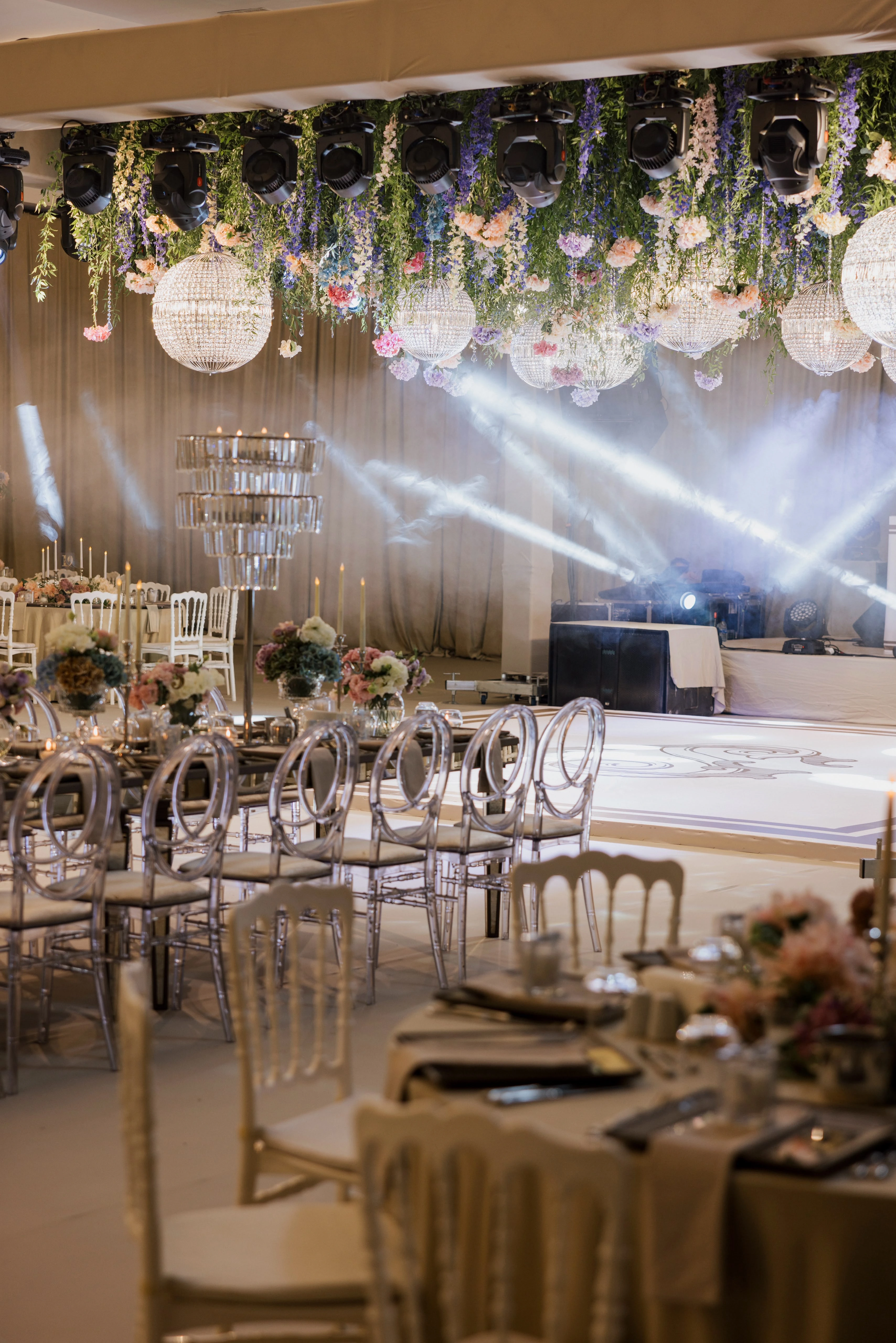 Round tables with silver chairs beneath stage light beams and a floral chandelier ceiling