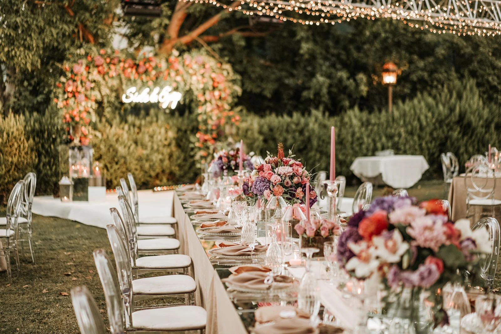 Long table with pink and purple flowers under string lights, a neon-lettered floral arch behind