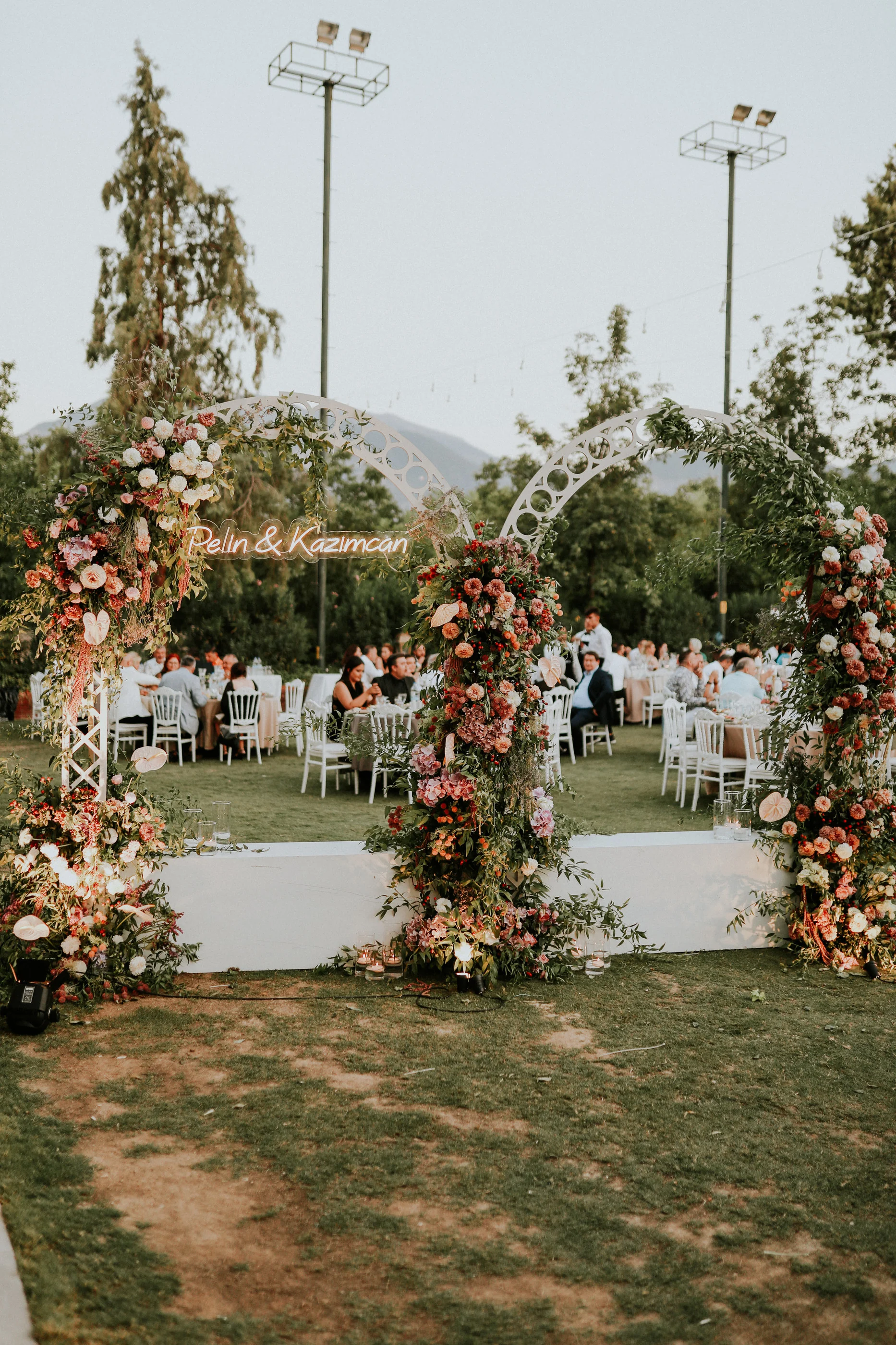 Colourful flower arches over a white platform with guests at round tables behind