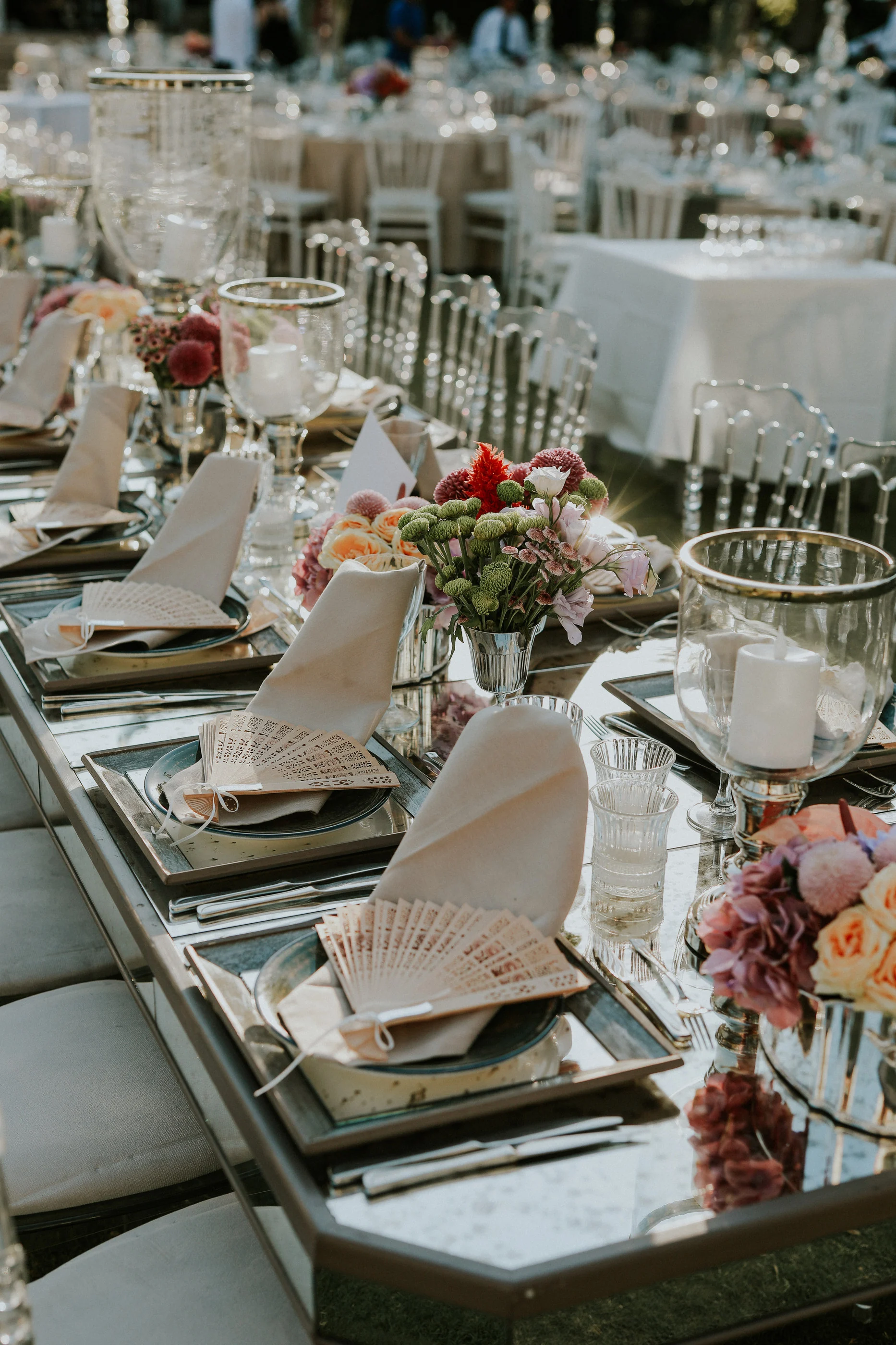 Fan-folded napkin, candle holder and flowers on a mirrored table, white-clothed tables behind
